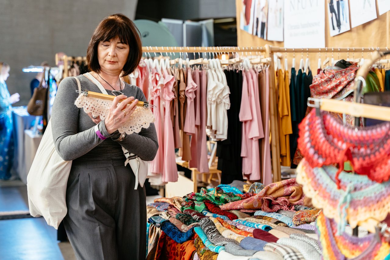Woman browsing handmade clothing and colorful textiles at an indoor artisan market.