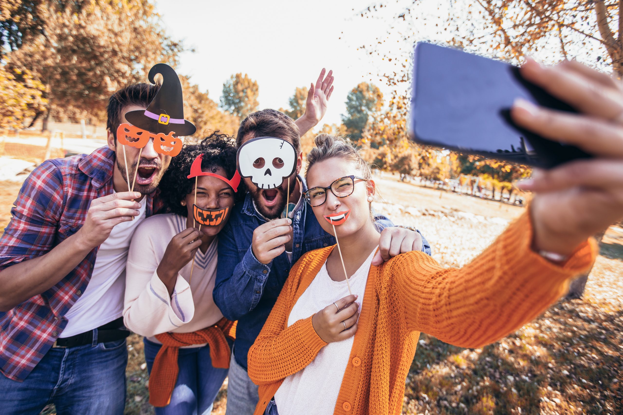 Group of friends taking a Halloween selfie outdoors with festive masks and autumn leaves in the background.